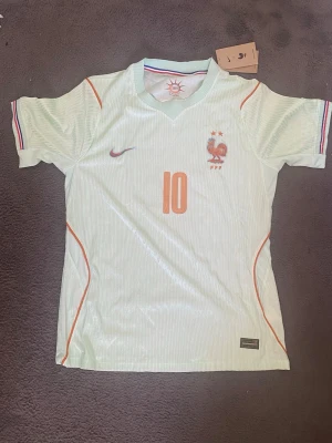 Frankrike fotbollströja Mbappé #10 - Ljusblå fotbollströja från Nike med Frankrikes landslagsemblem och nummer 10 på bröstet. På ryggen står det Mbappé och siffran 10 i orange. Tröjan har korta ärmar, tunna ränder och detaljer i orange samt trikolorfärgade muddar. Perfekt för fotbollsfans!
