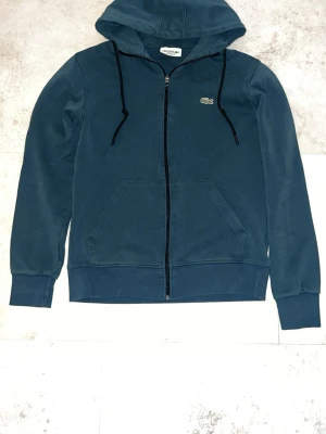 Blå zip-hoodie från Lacoste - Snygg blå hoodie från Lacoste med dragkedja, huva och svarta dragsnören. Klassisk krokodil-logga på bröstet och känguruficka framtill. Perfekt för en avslappnad och sportig stil.