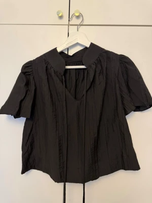 Svart blus med puffärm och knytband - Trendig svart blus med korta puffärmar och v-ringning. Blusen har en bred krage och dekorativa knytband framtill. Tillverkad i ett lätt, strukturerat tyg som ger en cool look. Perfekt att styla med jeans eller kjol.