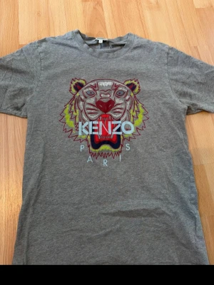 Grå Kenzo t-shirt med tigertryck - Säljer en grå t-shirt från Kenzo med det ikoniska tigertrycket i flera färger och texten 'KENZO PARIS' på bröstet. T-shirten har rund hals och korta ärmar, perfekt för en cool och avslappnad stil.