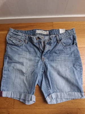 Ljusblå denimshorts med uppvik - Säljer ett par klassiska ljusblå jeansshorts i denim med fem fickor och uppvikta benslut. Shortsen har normal passform och bälteshällor i midjan. Perfekta för varma dagar och enkel att matcha med olika stilar.