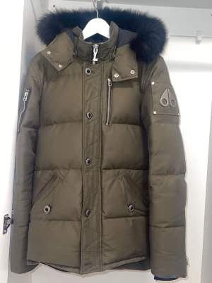 Olivgrön dunjacka med pälskant - Snygg olivgrön dunjacka med svart pälskant på huvan och flera fickor med dragkedja. Jackan har en modern passform, detaljer i metall och en patch på ärmen. Perfekt för kalla vinterdagar och riktigt varm tack vare dunfyllningen.