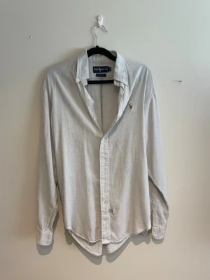 Vit skjorta från Ralph Lauren - Klassisk vit skjorta från Ralph Lauren med diskret rutigt mönster och broderad logga på bröstet. Skjortan har lång ärm, knappar framtill och en avslappnad passform. Perfekt till jeans eller chinos för en clean look.