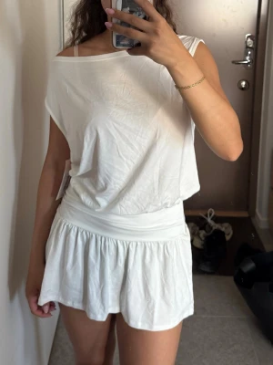 Vit kort klänning med offshoulder - Säljer en vit kort klänning med offshoulder-design och lös passform upptill. Kjoldelen har volang och klänningen är tillverkad i ett mjukt bomullsliknande material. Perfekt för varma dagar och enkel att styla med accessoarer.