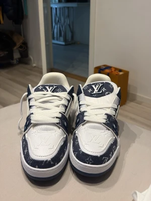 Louis Vuitton vita och blå sneakers - Snygga Louis Vuitton sneakers i vitt och marinblått med klassiskt LV-monogram på både tyg och läderdetaljer. Skorna har vita snören, robust sula och tydlig Vuitton-logga undertill. Perfekta för dig som vill sticka ut med exklusiv streetstyle.