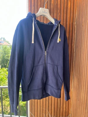 Mörkblå hoodie med dragkedja - Mörkblå hoodie med dragkedja och vita snören i huvan. Har två stora fickor framtill och en liten gul logga på bröstet. Tillverkad i mjukt bomullsmaterial och har ribbade muddar vid ärmar och nederkant. Perfekt för en avslappnad stil.