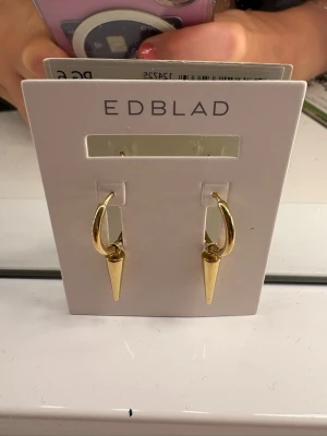 Guldpläterade örhängen från Edblad - Snygga guldpläterade örhängen från Edblad med rund ring och hängande konformad detalj. Stilren och modern design som ger en edgy touch till din look. Perfekta för dig som gillar minimalistiska men ändå statement smycken. Aldrig använd helt ny. Säljs i orginal förpackning. Aldrig använd pga att jag är silver och inte guld.