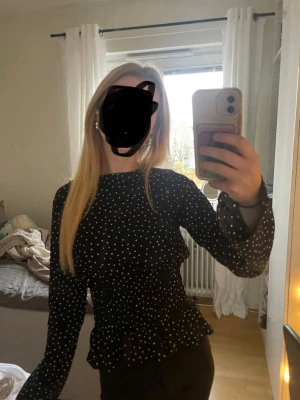 Svart prickig blus med volang - Trendig Snygg svart blus med vita prickar och volangdetaljer i midjan. Blusen har långärmade ärmar med lätt transparent tyg och smockad midja som ger en feminin siluett. Perfekt att styla med jeans eller kjol för en trendig look.