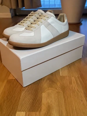 Maison Margiela Gats - Stilrena sneakers från Maison Margiela i vitt och beige läder och mocka. Skorna har klassisk låg silhuett, beige snörning och gummisula. Perfekta för dig som gillar minimalistisk och tidlös design.