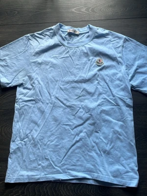 Ljusblå Moncler t-shirt i bomull - Moncler T-shirt – Ljusblå – Storlek XS  Säljer en stilren Moncler t-shirt i ljusblå färg och storlek XS. Tröjan är i 9/10 skick och mycket väl omhändertagen, med endast minimala tecken på användning.  Detaljer:  * Storlek: XS * Färg: Ljusblå * Skick: 9/10 * Mjuk och bekväm bomull *Skickar inom 24 timmar