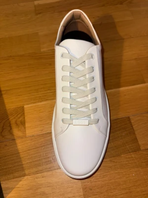 Vita sneakers från BOSS (helt nya) - Stilrena vita sneakers från BOSS i slät läderlook. Klassisk låg modell med vita snören och diskret logga på plösen. Snygg och minimalistisk design som passar till det mesta. Kommer i originalkartong. Helt nya (ny pris 4000kr) mitt pris 1200
