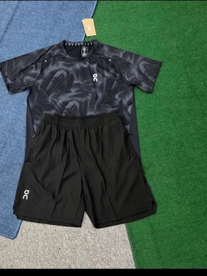 Svart träningsset från On - Snyggt träningsset från On med en svart t-shirt i abstrakt mönster och matchande svarta shorts. T-shirten har rund hals och korta ärmar, medan shortsen har elastisk midja och sidofickor. Perfekt för gymmet eller löprundan.