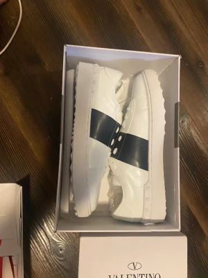 Vita sneakers med svart band Valentino - Snygga vita sneakers från Valentino med ett brett svart band över mitten och dekorativa nitar. Skorna har en chunky sula och är tillverkade i slätt läder. Perfekta för dig som vill ha en clean men ändå lyxig look. Jag köpte fel size och det var inga retur tillåtna det stog att de var äkta och de känns som  de men jag är inte helt säker på det.