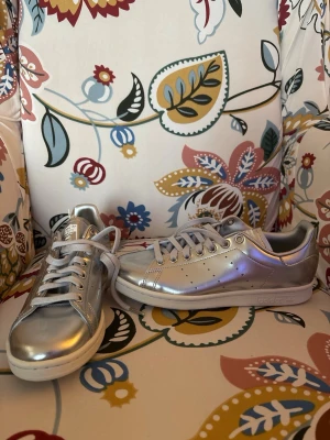 Silvriga sneakers med metallicfinish - Snygga sneakers i metallic-silver från Adidas med klassisk låg profil och vita sulor. Skorna har snörning och diskreta perforeringar på sidorna. Perfekta för dig som vill sticka ut med en futuristisk vibe och cool look.