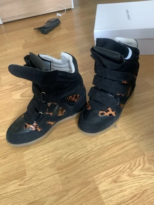 Isabel marant - Unika svarta sneakers med högt skaft och leopardmönstrade detaljer. Skorna har flera kardborreband och är tillverkade i mockaliknande material med vita vadderade insidor. Perfekt för dig som vill sticka ut med din stil.