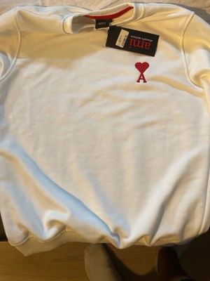 Vit sweatshirt från Ami Paris - Säljer en stilren vit sweatshirt från Ami Paris med det ikoniska röda hjärtat och bokstaven A broderat på bröstet. Tröjan har rund halsringning och ribbade muddar. Perfekt för en clean och trendig look.
