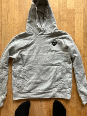 Grå hoodie med svart hjärta - Säljer en grå hoodie med huva och magficka. På bröstet sitter en svart hjärtdetalj med ögon, vilket ger en unik och cool look. Hoodien har dragsko i huvan och är tillverkad i mjukt sweatshirtmaterial. Perfekt för dig som gillar streetwear och enkel stil.