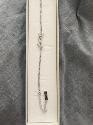 Silverfärgat armband från Saint Laurenn - Snyggt armband i silverfärg från Saint Laurent med den ikoniska YSL-logotypen som hänge. Kedjan är tunn och har en liten bricka med texten 'Saint Laurent Paris' vid låset. Perfekt accessoar för att lyfta din outfit.