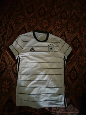 Vit Adidas Tyskland fotbollströja - Vit fotbollströja från Adidas med svarta horisontella ränder, Tysklands landslagslogga och fyra stjärnor på bröstet. Svart krage och ärmslut med tyska flaggans färger. Perfekt för fotbollsträning eller match.
