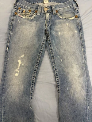 Ljusblå slitna jeans True Religion - Säljer ett par ljusblå jeans från True Religion med slitningar och distressed detaljer. Klassisk femficksmodell med kontrastsömmar och stora bakfickor med lock och knappar. Jeansen har en avslappnad passform och är tillverkade i bomull.
