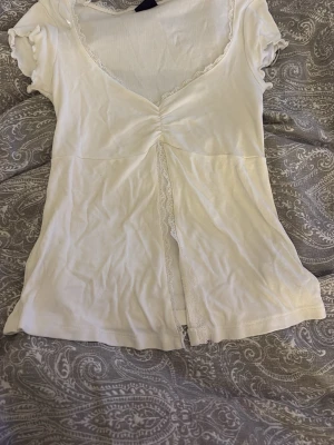 Vit topp med spets och rynkdetalj - Söt vit topp med v-ringning, rynkdetalj framtill och spetskant längs halsringningen och mittsömmen. Kortärmad och lätt figursydd modell som ger en feminin look. Perfekt att styla med jeans eller kjol.