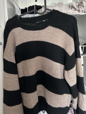 Stickad tröja med breda ränder - Säljer en oversized stickad tröja med breda svarta och beige ränder. Tröjan har rund halsringning och långa ärmar. Perfekt för lager på lager och chill stil. Materialet känns mjukt och bekvämt.