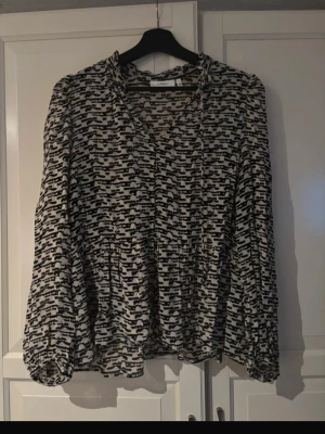 Mönstrad blus från InWear - Snygg blus från InWear i svartvitt abstrakt mönster. Blusen har v-ringning med liten volangkant och långa ärmar med resår vid handlederna. Luftig och lätt i materialet, perfekt för en trendig look. Storlek 38.