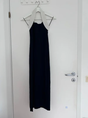 Svart långklänning med vit krage - Stilren svart långklänning med bred vit krage och tunna axelband. Klänningen har en rak siluett och är golvlång, vilket ger en elegant look. Perfekt för dig som vill ha en klassisk och tidlös stil.