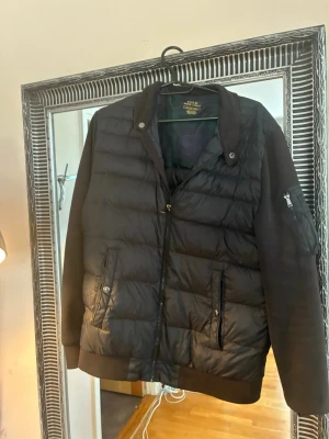 Svart cardigan från Polo Ralph Lauren - Säljer en svart pufferjacka från Polo Ralph Lauren med quiltad front, mjuka ärmar och ribbade muddar. Jackan har två fickor med knappstängning och en ärmficka med dragkedja. Insidan har rutigt foder för extra stilpoäng.