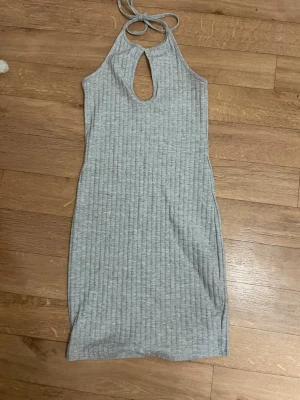 Grå ribbad halterneckklänning - Snygg grå ribbad klänning med halterneck och knyt i nacken. Klänningen har en liten öppning framtill och är ärmlös. Perfekt för en trendig och avslappnad look.