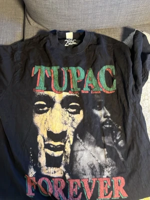 Svart Tupac Forever t-shirt - Säljer en svart t-shirt med stort Tupac Forever-tryck fram och bak. Framsidan har Tupacs ansikte och texten 'TUPAC FOREVER' i rött och grönt, medan baksidan har en bild av Tupacs rygg och texten 'The World Against'. Perfekt för dig som gillar hiphop och streetwear. Köpt ifrån jumpyard! Hör gärna av dig vid fråga eller fundering!!!