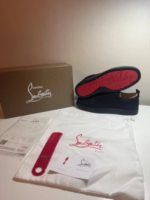 Svarta Louboutin sneakers med nitar - Snygga svarta sneakers från Christian Louboutin med klassisk röd sula och coola nitar på tån. Skorna har lågt skaft och är tillverkade i mocka med läderdetaljer. Perfekta för dig som vill sticka ut med exklusiv design.