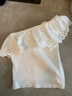 Vit offshoulder topp med volang - Superfin vit offshoulder topp med bred volangkant och broderade blommor. Toppen är ribbad och har en romantisk känsla, perfekt till sommaren. Sitter snyggt och är lätt att matcha med både jeans och kjol.