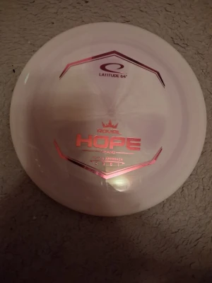 Vit discgolf disc Latitude 64 Hope - Säljer en vit discgolf disc från Latitude 64, modellen Royal Hope. Discen har rosa metallic-tryck och är rund med slät yta, perfekt för putt och approach. Snygg design och skön att kasta för både nybörjare och erfarna spelare. Helt ny!