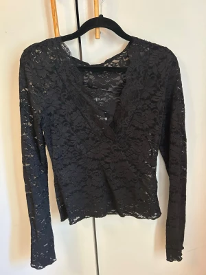 Svart spetsblus med v-ringning - Säljer en svart långärmad blus i spets med v-ringad omlottfront. Blusen är genombruten och har en feminin look med fina spetsdetaljer. 