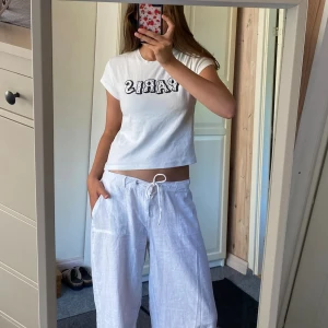 Vit t-shirt med PARIS-tryck - Säljer en vit t-shirt med svart text där det står PARIS på bröstet. T-shirten har korta ärmar, rund halsringning och är i en croppad modell. Perfekt till jeans eller shorts för en avslappnad look.