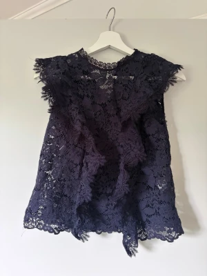 Mörkblå spetsblus från Zara - Supersnygg mörkblå blus från Zara i genombruten spets med blommönster. Ärmlös modell med volangdetaljer vid axlarna och fin rund halsringning. Perfekt att styla med jeans eller kjol för en trendig look.