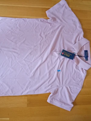 Ljusrosa pikétröja från Polo Ralph Lauren - Snygg ljusrosa pikétröja från Polo Ralph Lauren med klassisk krage och korta ärmar. Tröjan har en blå broderad logga på bröstet och är tillverkad i mjuk bomull. Perfekt för en clean och stilren look.