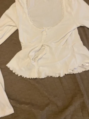 Vit långärmad blus med volangkant - Söt vit blus med lång ärm, djup rundringning och dekorativ volangkant nertill. Blusen har knyt framtill och små spetsdetaljer längs kanterna. Perfekt för en trendig och feminin look.