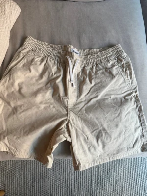 Beige shorts från Weekday med snörning - Snygga beige shorts från Weekday med elastisk midja och vit snörning framtill. Tillverkade i mjuk bomull, perfekta för varma dagar. Två sidofickor ger en avslappnad vibe och enkel stil.