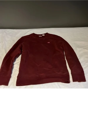 Vinröd sweatshirt med rund hals - En enkel och stilren vinröd sweatshirt med rund halsringning och långa ärmar. Tröjan har en liten diskret logga på bröstet och ribbade muddar vid ärmslut och nederkant. Perfekt för en avslappnad look.