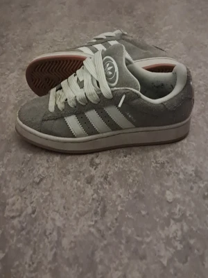 Gråa sneakers med vita ränder - Snygga gråa sneakers från Adidas med klassiska vita ränder på sidan och vit sula. Skorna har en ovandel i mocka och gummisula med fiskbensmönster undertill. Perfekta för dig som gillar en clean och sportig stil.
