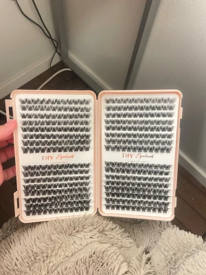 DIY Eyelash fransar i mixad längd - Säljer ett kit med svarta individuella ögonfransar i olika längder, perfekta för att skapa en personlig franslook. Fransarna kommer i en ljusrosa ask med texten 'DIY Eyelash' och är gjorda av syntetmaterial. Perfekt för dig som vill fixa fransarna själv hemma.