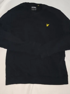 Svart sweatshirt från Lyle & Scott - Klassisk svart sweatshirt från Lyle & Scott med rund halsringning och gul broderad logga på bröstet. Tröjan har långa ärmar och ribbade muddar vid ärmslut och nederkant. Perfekt för en clean och stilren look.
