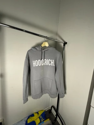 Grå HOODRICH hoodie med tryck - Snygg grå hoodie från HOODRICH med stor vit logga på bröstet. Klassisk modell med huva och dragsnören samt en stor magficka. Perfekt för en chill och avslappnad stil. Tillverkad i mjukt material för extra komfort.