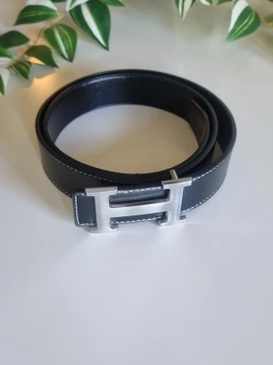 Svart bälte - Snyggt svart bälte i skinn med ett stort, silverfärgat H-format spänne. Bältet har vita kontrastsömmar längs kanterna som ger en clean och modern look. Perfekt accessoar för att lyfta vilken outfit som helst.