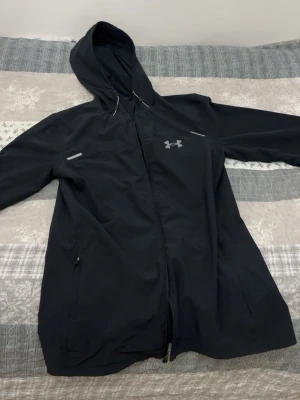  Under Armour jacka - Snygg svart vindjacka från Under Armour med huva och dragkedja framtill. Jackan har reflekterande detaljer, två sidofickor med dragkedja och logga på bröstet samt stor logga på ryggen. Tillverkad i lätt och tåligt material, perfekt för blåsiga dagar. Den är S passas M