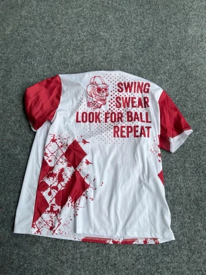 Vit och röd golftröja med tryck - Unik golftröja i vitt och rött med coolt dödskalletryck och texten 'SWING SWEAR LOOK FOR BALL REPEAT' på ryggen. Framsidan har ett stort motiv och ordet 'GOLF'. Tröjan har korta ärmar och är tillverkad i ett lätt, funktionsmaterial som passar perfekt för sport.