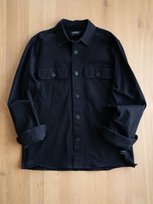 Svart overshirt från Gabba - Stilren svart overshirt från Gabba med klassisk krage och knäppning framtill. Två bröstfickor med lock och knappar ger en schysst look. Tillverkad i ett mjukt, slitstarkt material som känns bekvämt. Perfekt att bära öppen eller stängd över en t-shirt.