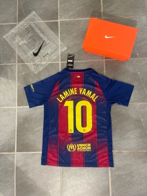 FC Barcelona fotbollströja Lamine Yamal - Säljer en officiell FC Barcelona fotbollströja från Nike med Lamine Yamal och nummer 10 på ryggen. Tröjan har blå och röda vertikala ränder, klubbmärke och Spotify-logga framtill samt UNHCR-logga nedtill. Korta ärmar och sportig passform, perfekt för fotbollsfans.